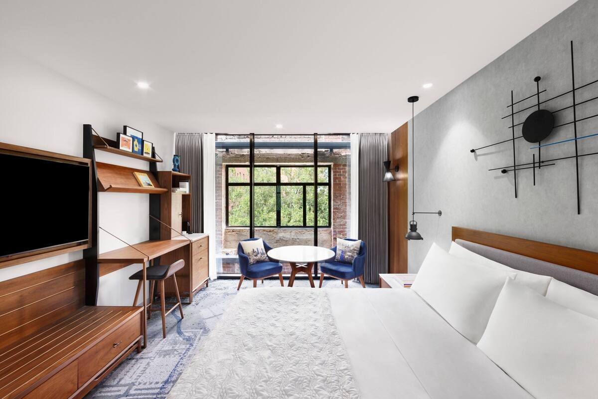A modern hotel room with a large bed, a TV, a desk with a chair, a round table with two chairs, and a large window with a view of greenery.
