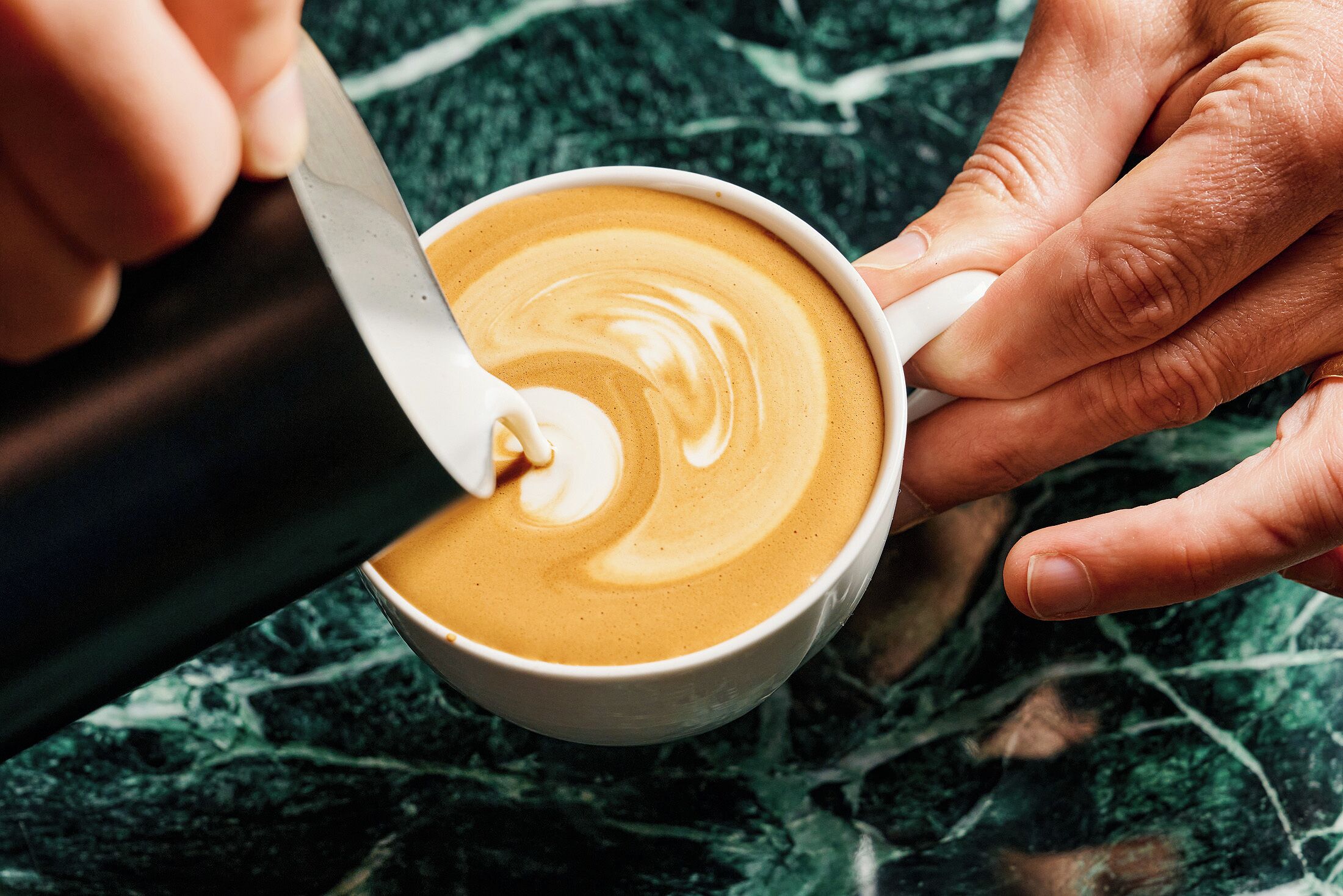 Een detailfoto van handen die een kop koffie met een schuimige laag roeren met een lepel.