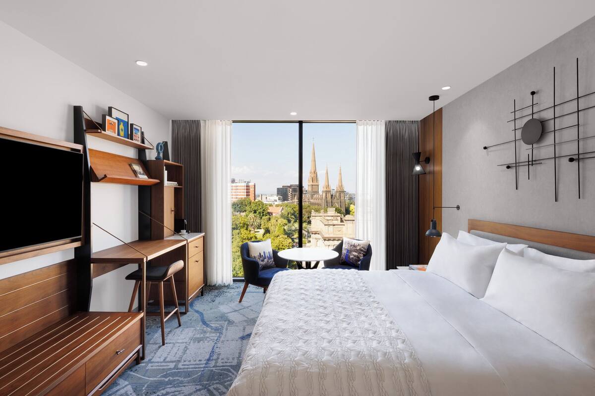 A hotel room with a bed, desk, chair, TV, and a view of a cityscape.