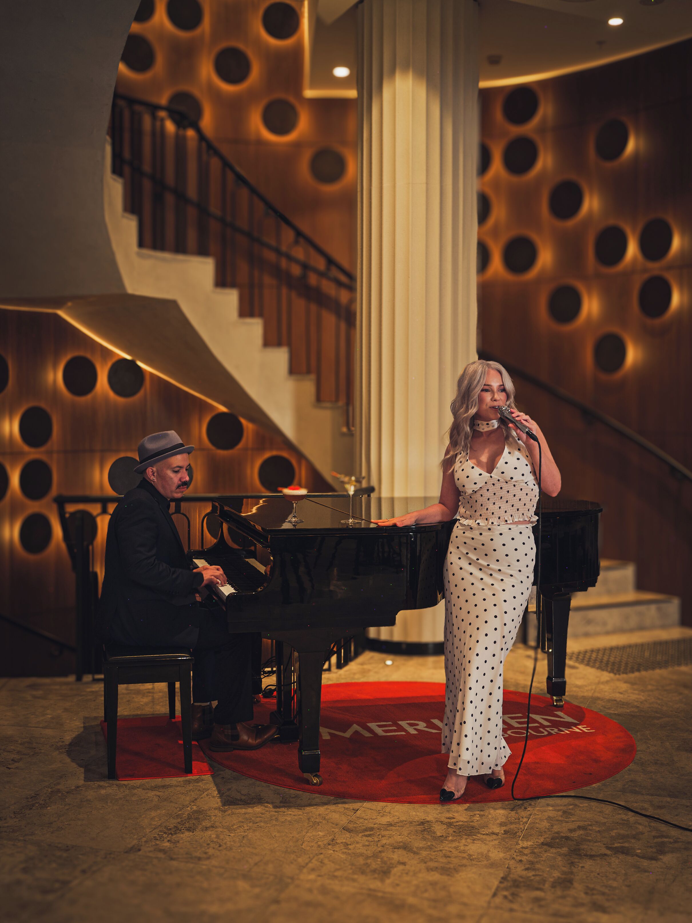 Un homme joue du piano et une femme chante sur un tapis rouge dans une pièce dotée d’un escalier et dont les murs sont de forme circulaire.
