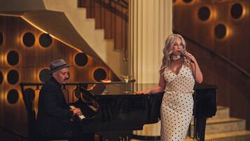 A man playing a piano and a woman singing on a red carpet in a room with a staircase and circular wall design.
