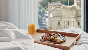 A breakfast tray with waffles, a croissant, and a glass of orange juice on a bed with white linens, overlooking a cityscape with historic buildings. - Le Méridien Melbourne (Melbourne)