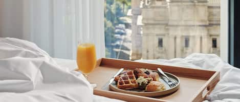A breakfast tray with waffles, a croissant, and a glass of orange juice on a bed with white linens, overlooking a cityscape with historic buildings.
