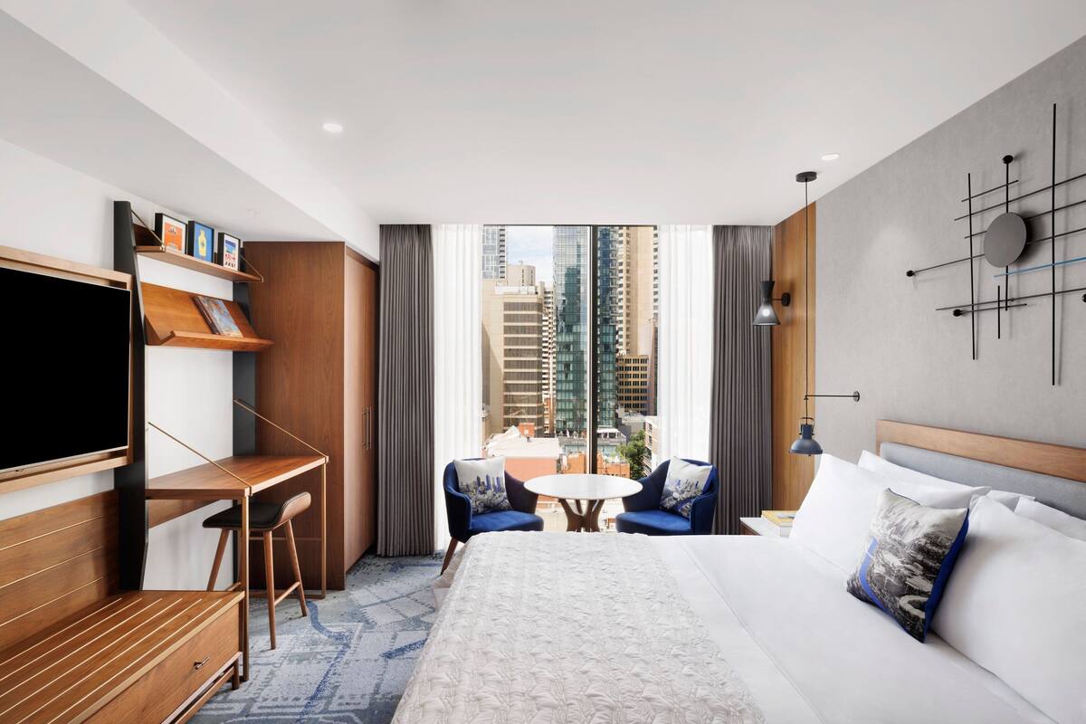 A hotel room with a bed, a desk, a TV, a chair, a small table, and a view of the city.