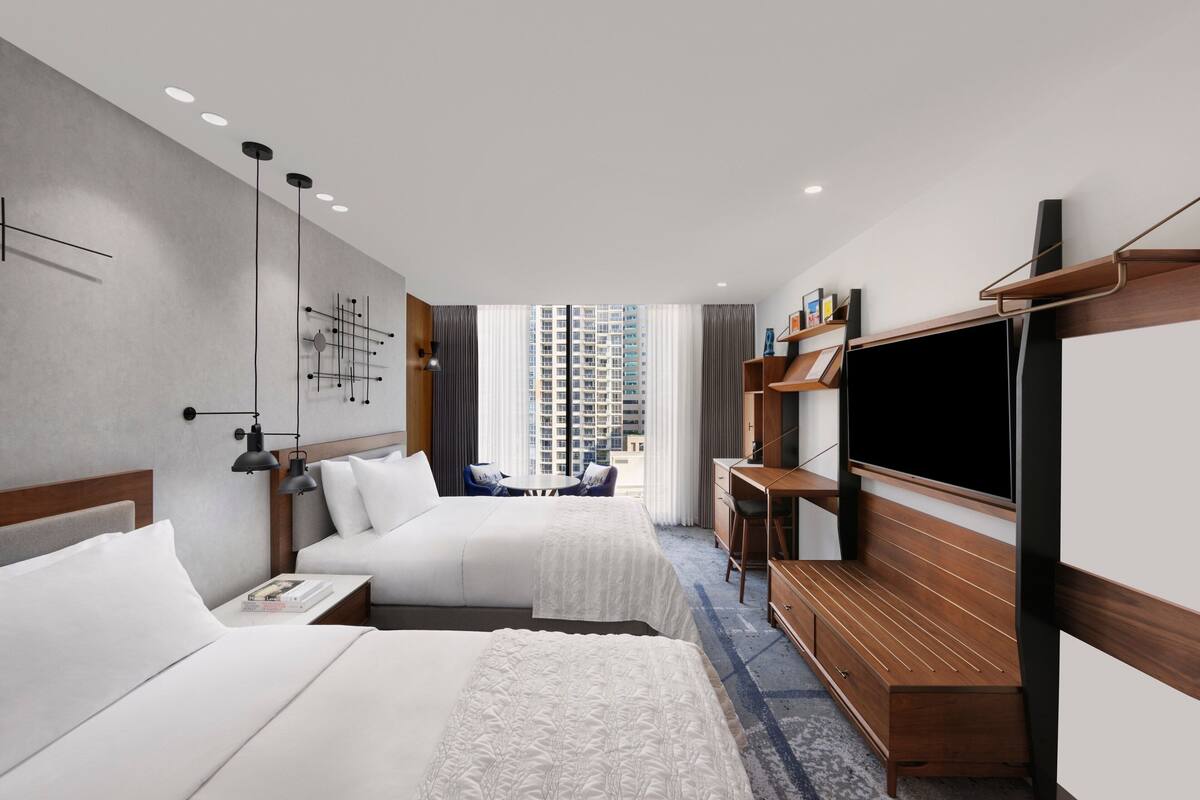A hotel room with two beds, a TV, a desk, and a view of a cityscape.