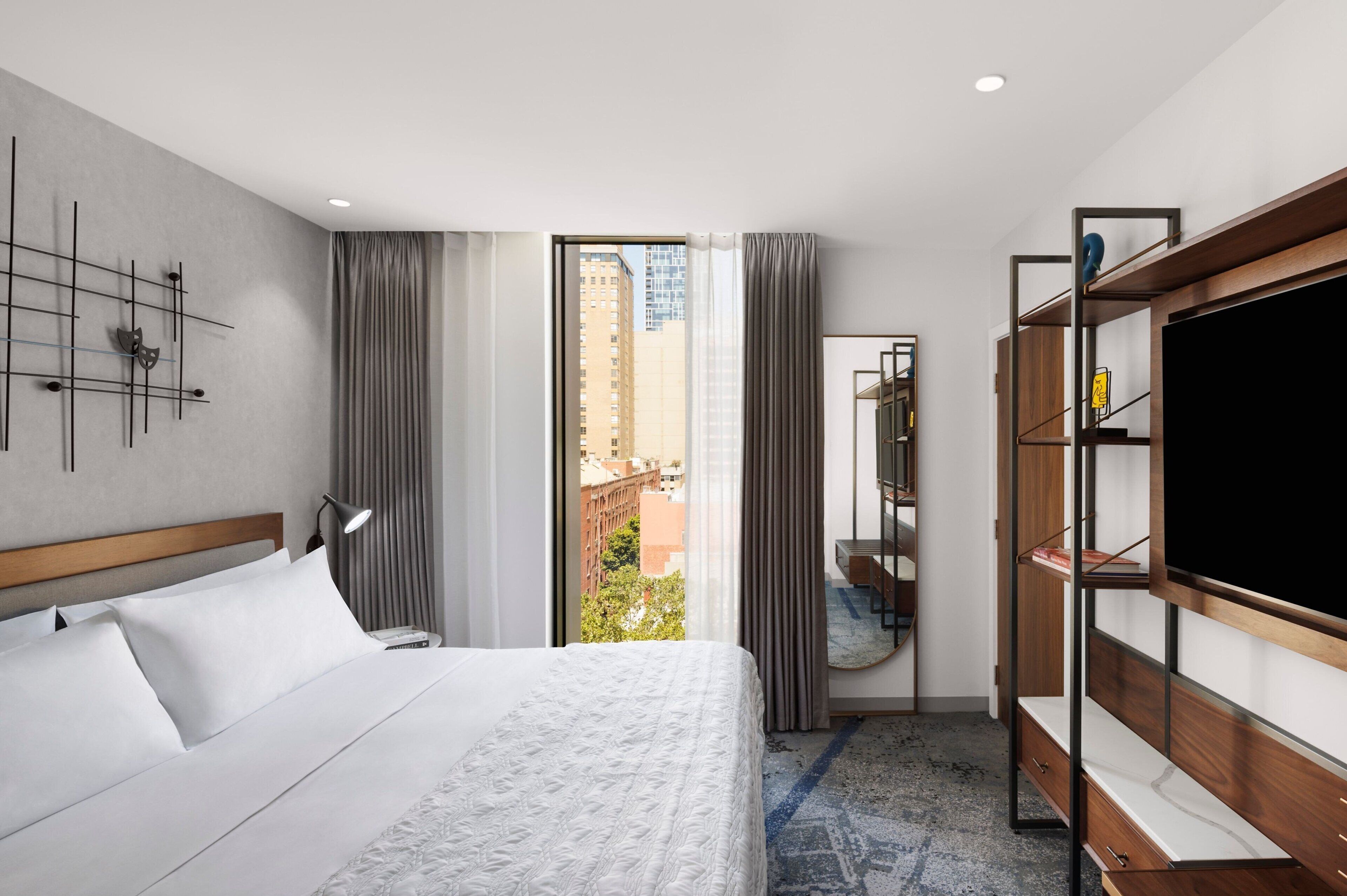 Habitación de hotel con cama, televisor, armario y vistas a edificios a través del ventanal.