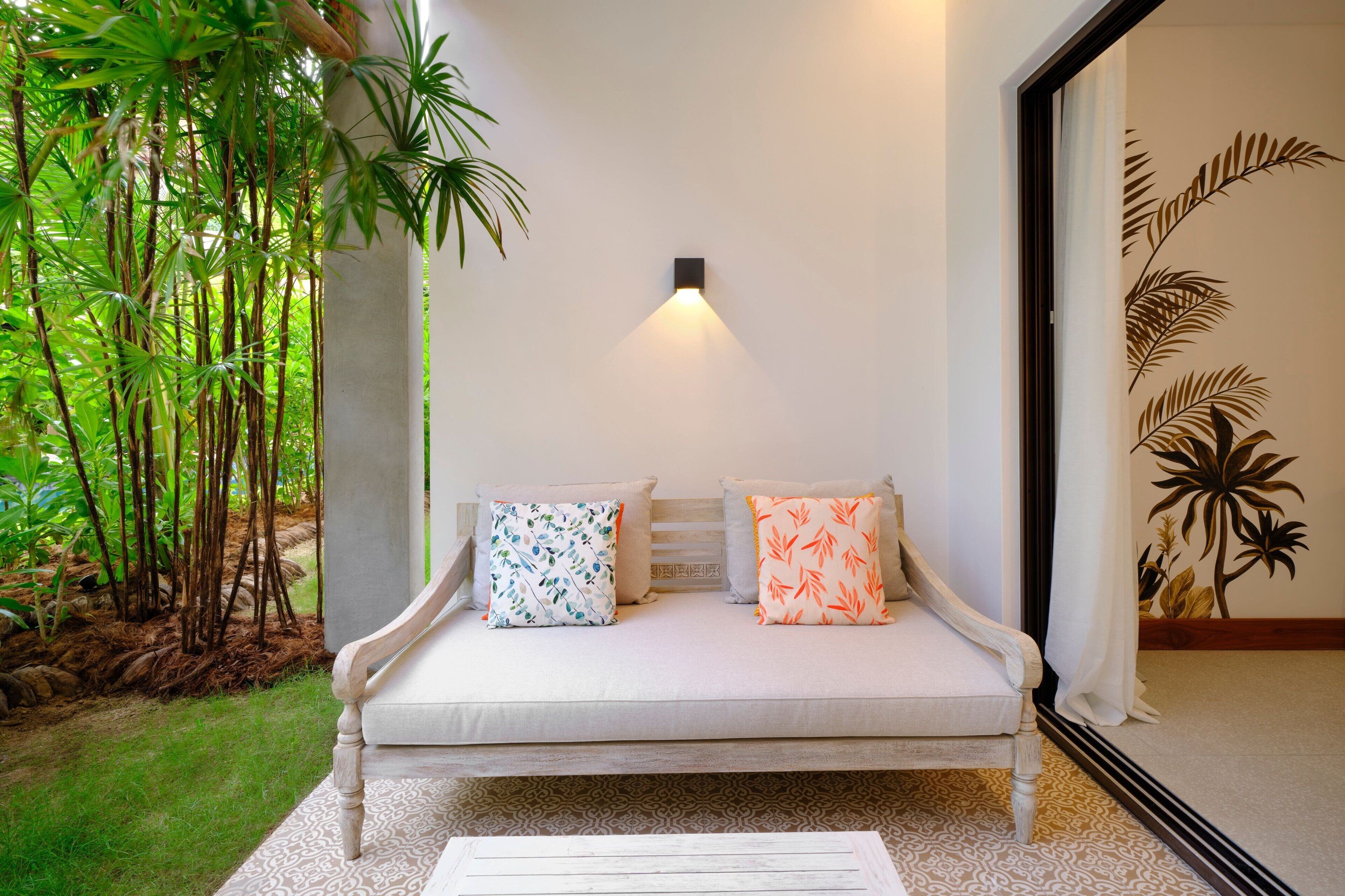 A white sofa with patterned cushions, a small table, and a wall-mounted light fixture in a room with a large mirror and a decorative wall panel.