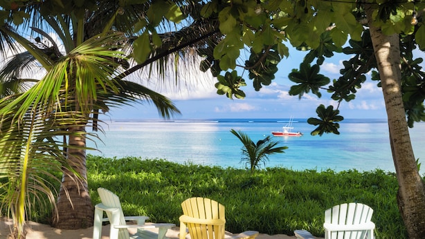 Ein Sitzbereich direkt am Strand mit Stühlen und einem Tisch, mit Blick auf ein ruhiges Meer und ein Boot in der Ferne.
