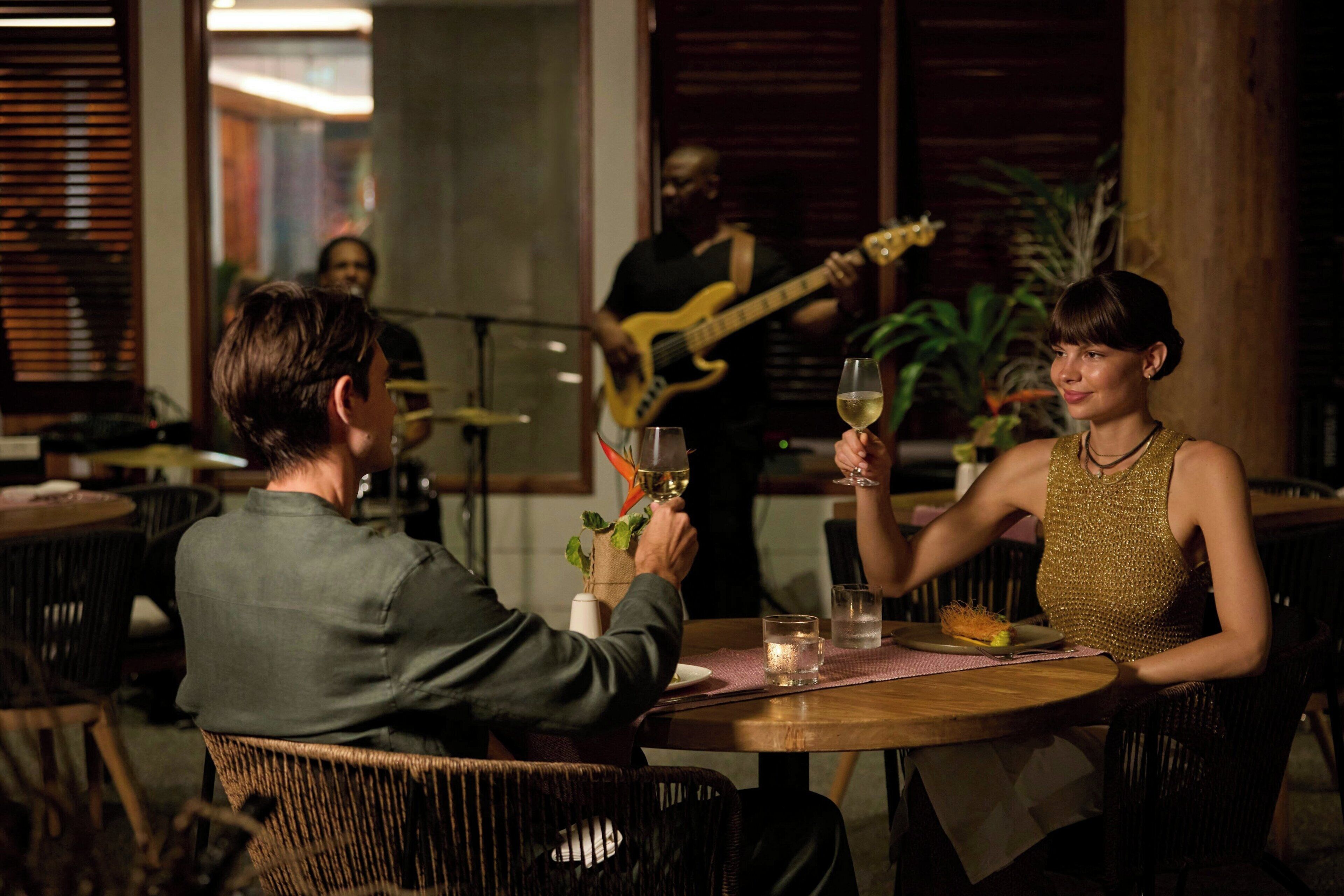 A couple seated at a wooden table with a glass of wine, a small plant, and a menu. A musician plays a guitar in the background.