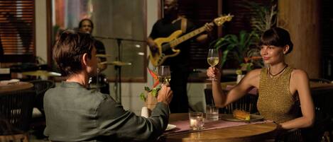 A couple seated at a wooden table with a glass of wine, a small plant, and a menu. A musician plays a guitar in the background.