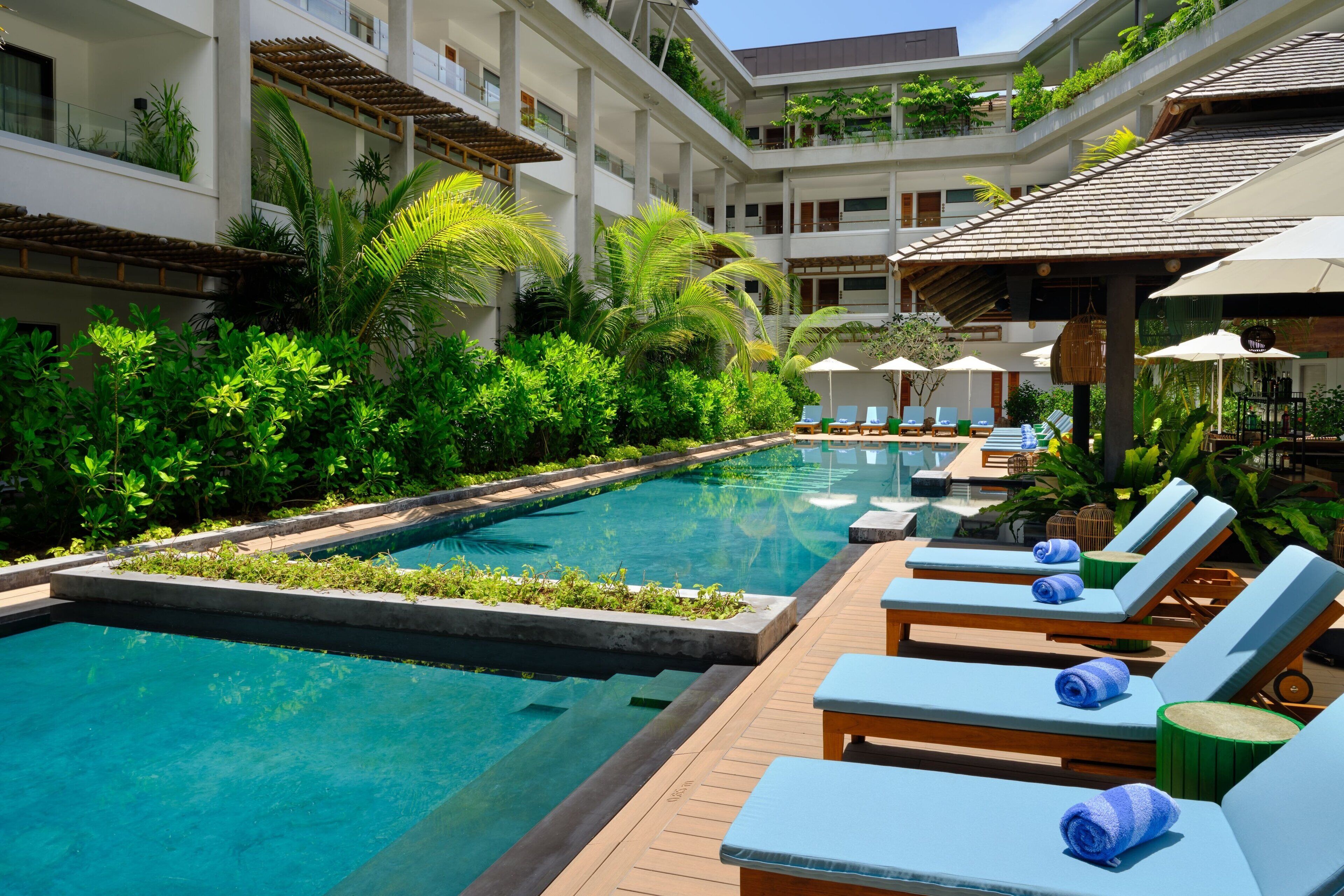 A pool area with lounge chairs and umbrellas, surrounded by lush greenery and a multi-story building with balconies.