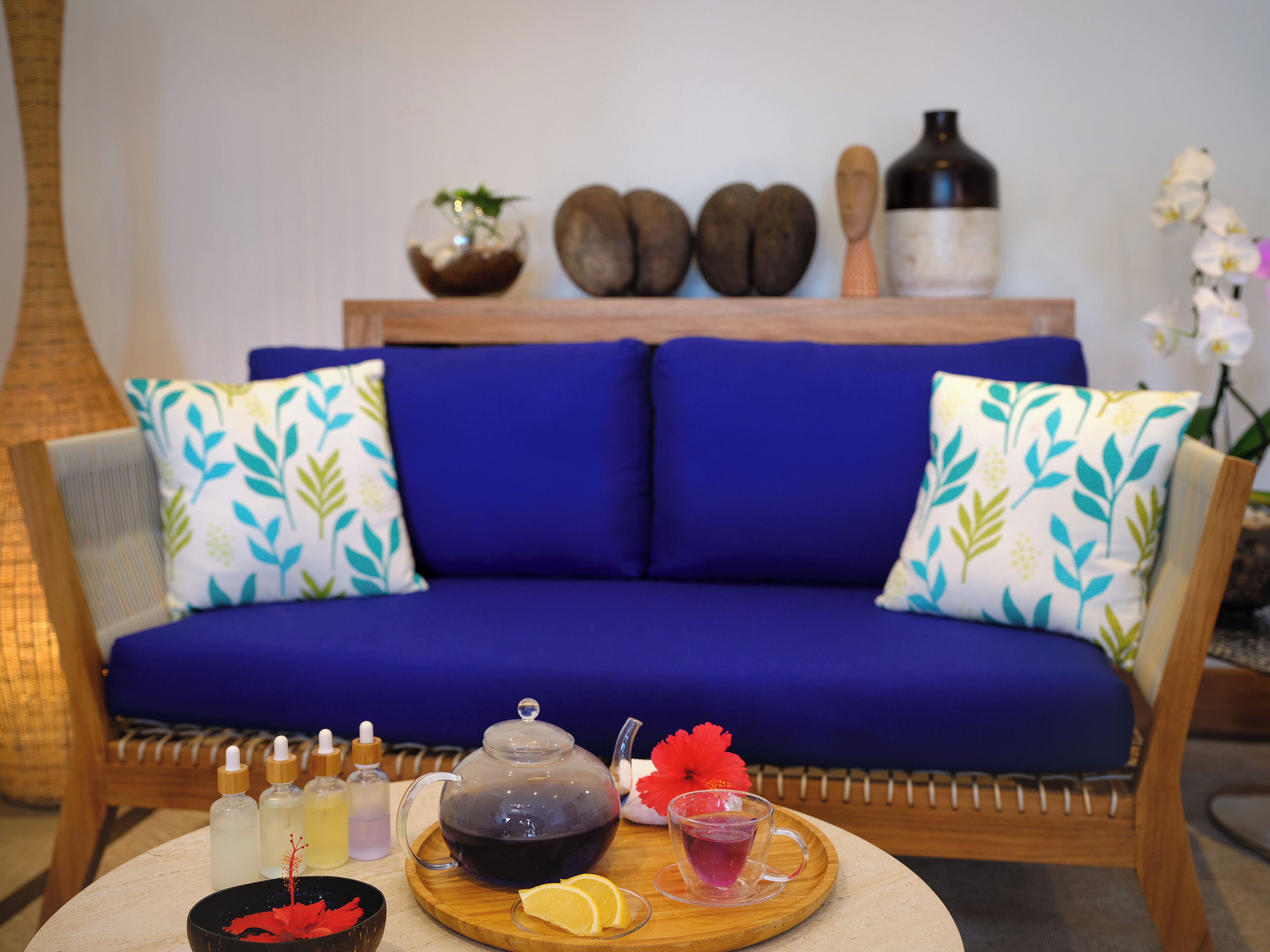 Un sofá azul con cojines estampados, una bandeja de madera con tetera, tazas de té y botellas, y objetos decorativos en una pared de fondo.