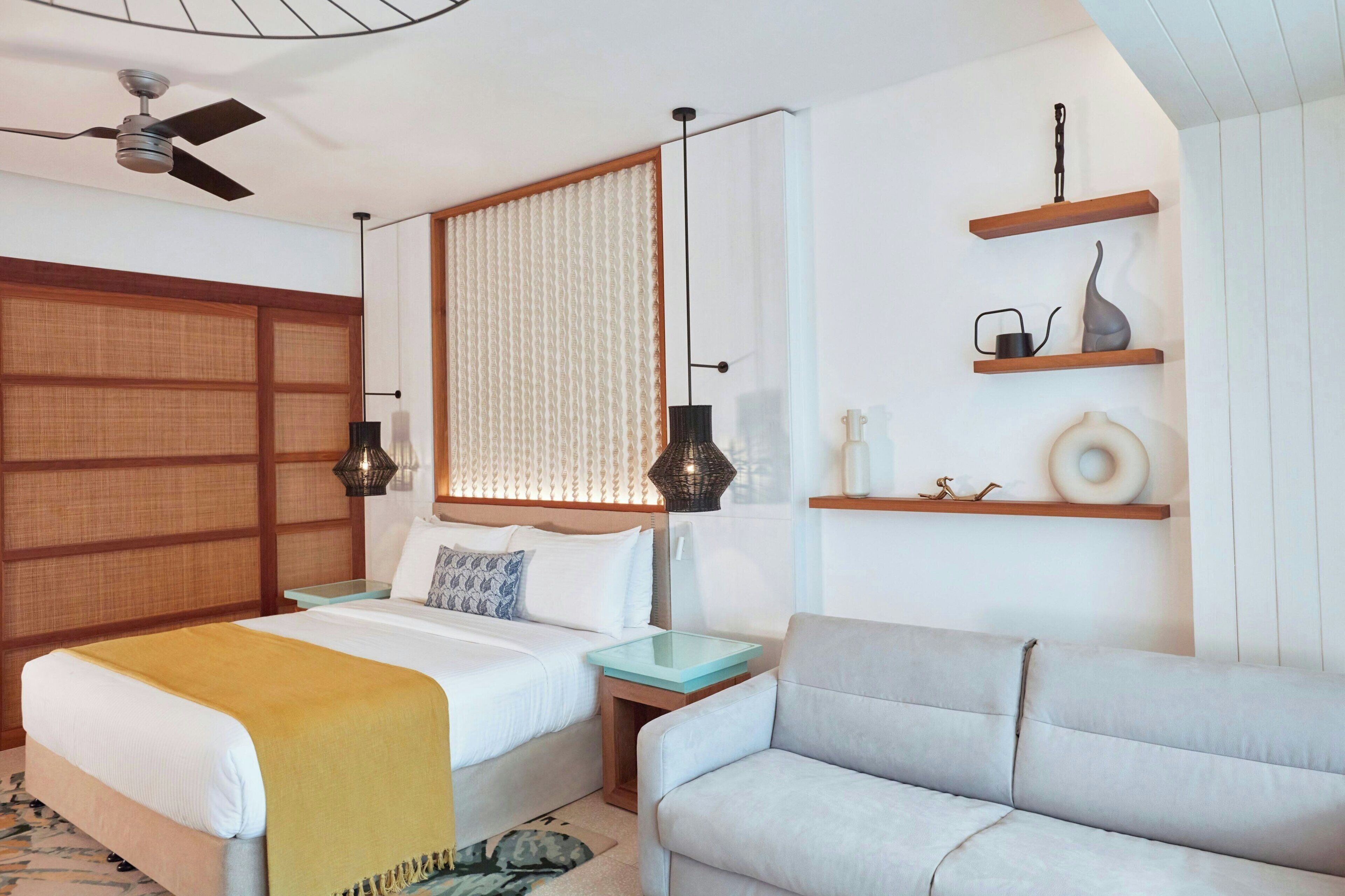 A hotel room with a bed, a sofa, a ceiling fan, a window with blinds, and a shelf with decorative items.