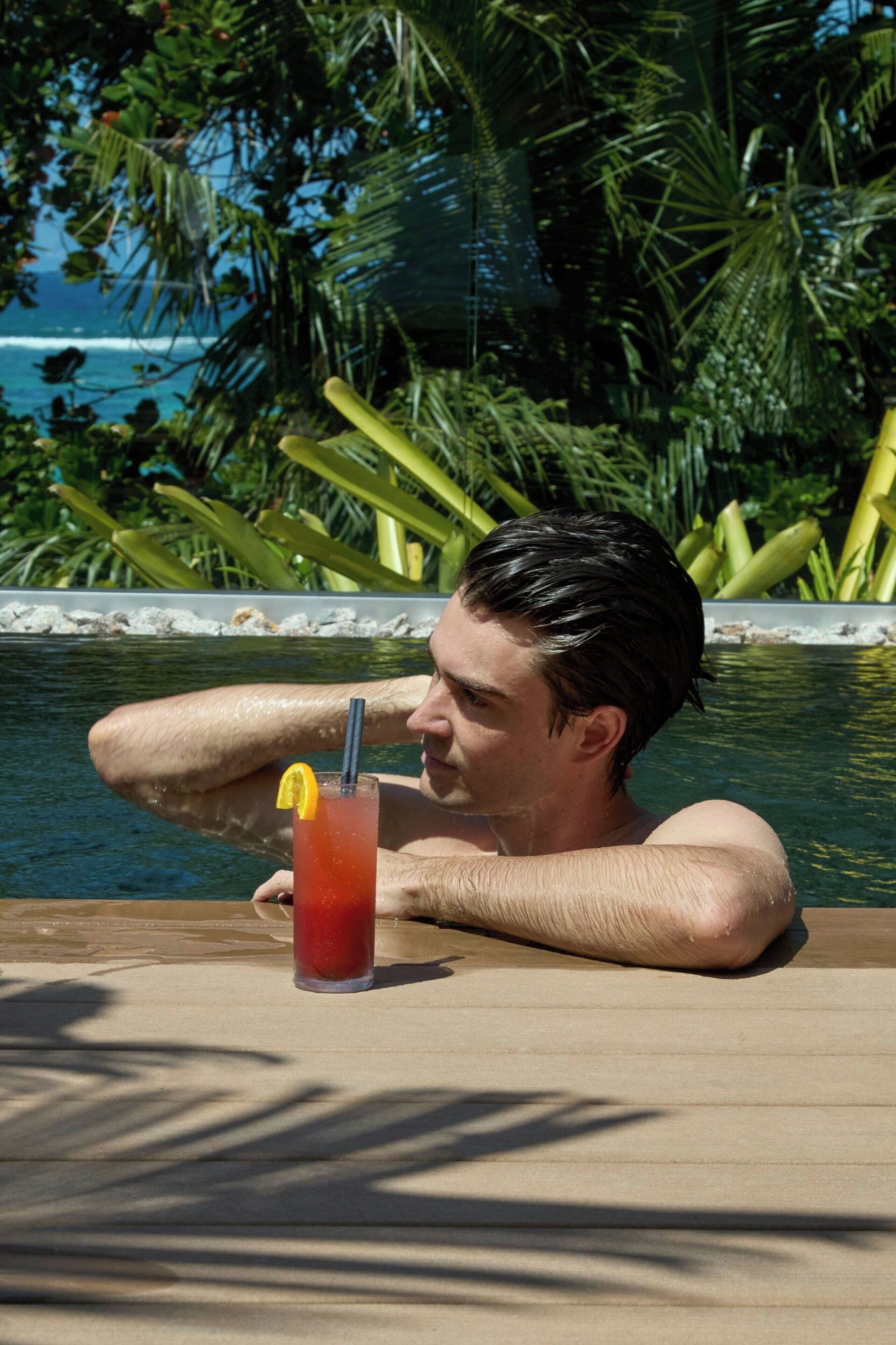 A man relaxing by a pool with a drink, surrounded by lush greenery and a view of the sea.