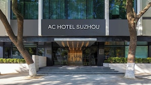 La entrada del Hotel AC Suzhou, con fachada moderna, amplios ventanales y una entrada acogedora con escalones y luces iluminadas.
