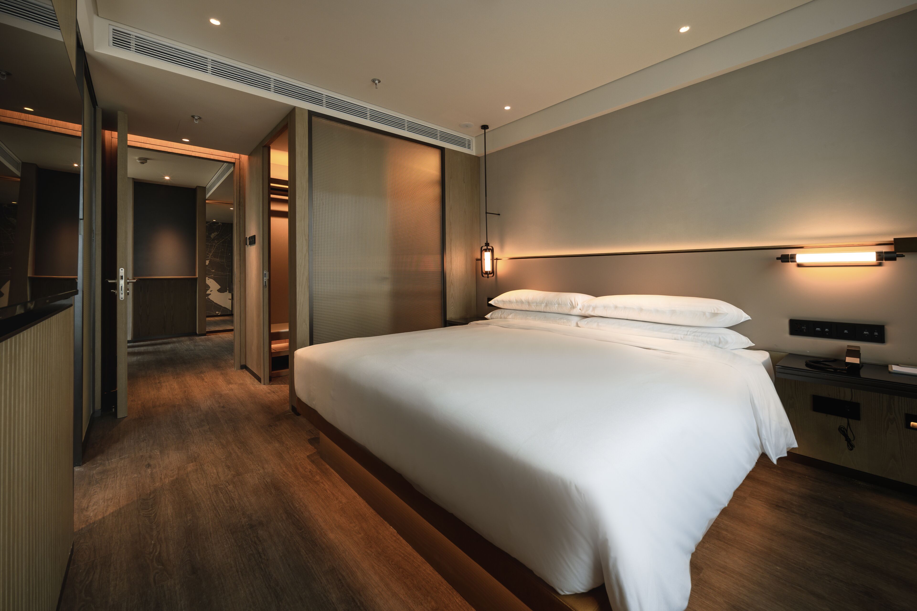 A modern hotel room with a large bed, a bedside table, a built-in TV, and a walk-in closet.