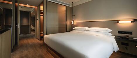 A modern hotel room with a large bed, a bedside table, a built-in TV, and a walk-in closet.