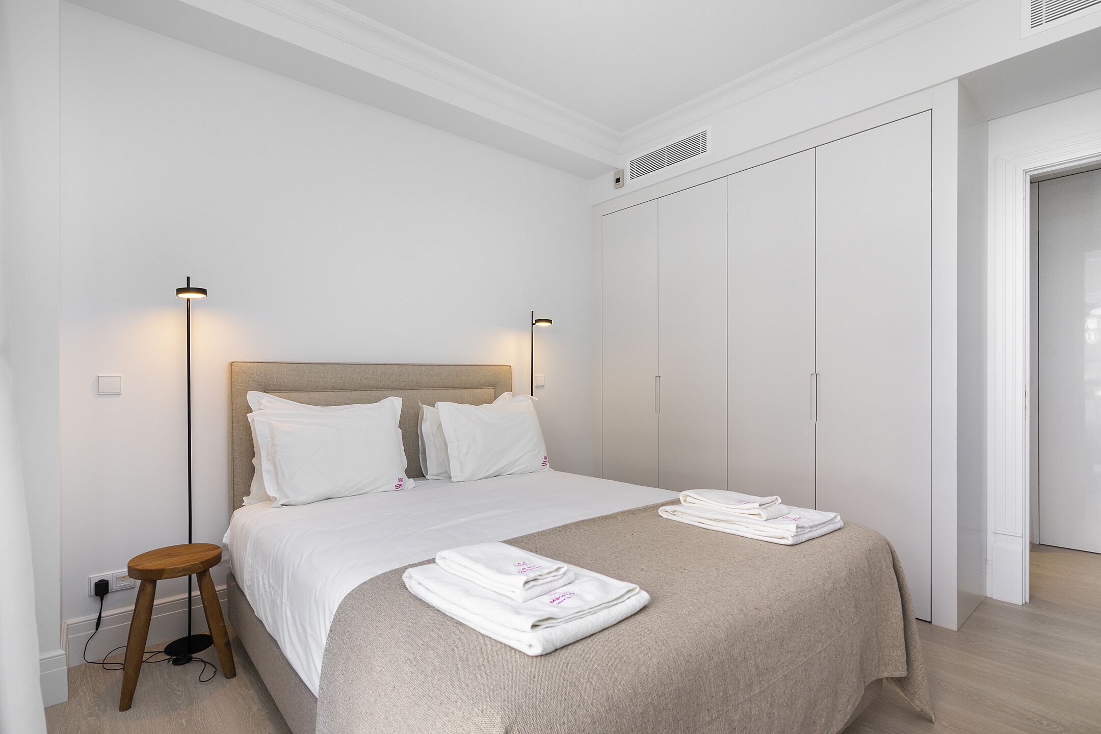 A bedroom with a bed, bedside lamps, a wooden stool, and neatly folded towels on the bed.