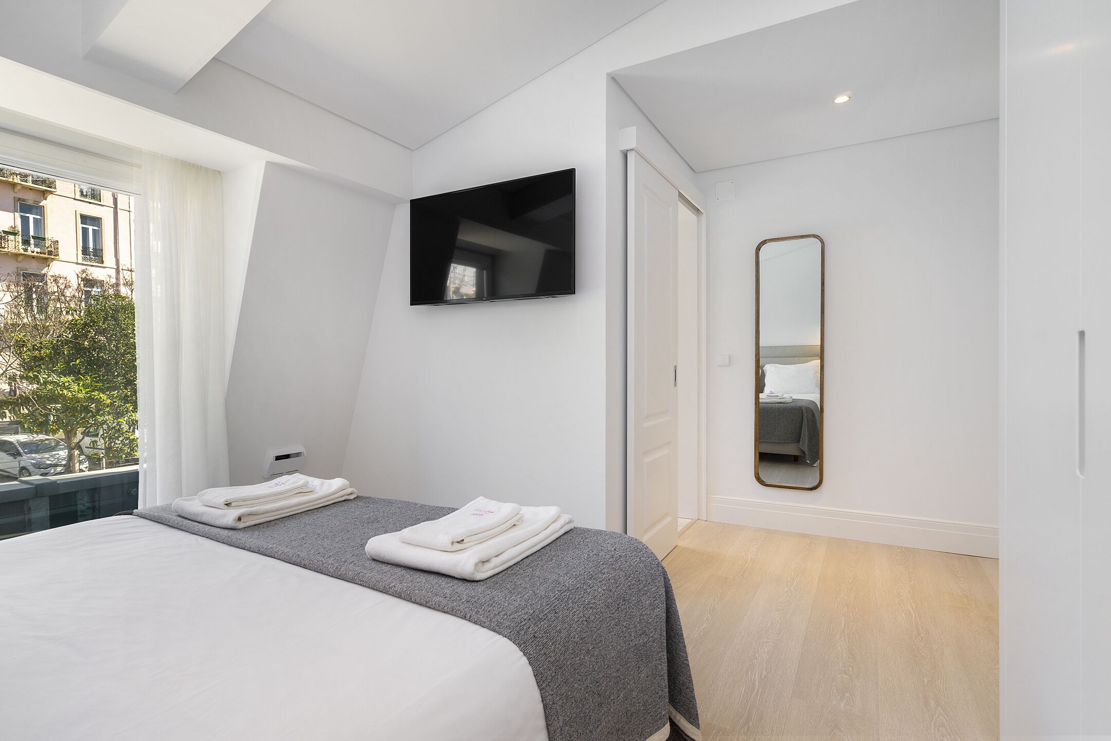 A modern bedroom with a large bed, a flat-screen TV, a mirror, and a view of the outside.