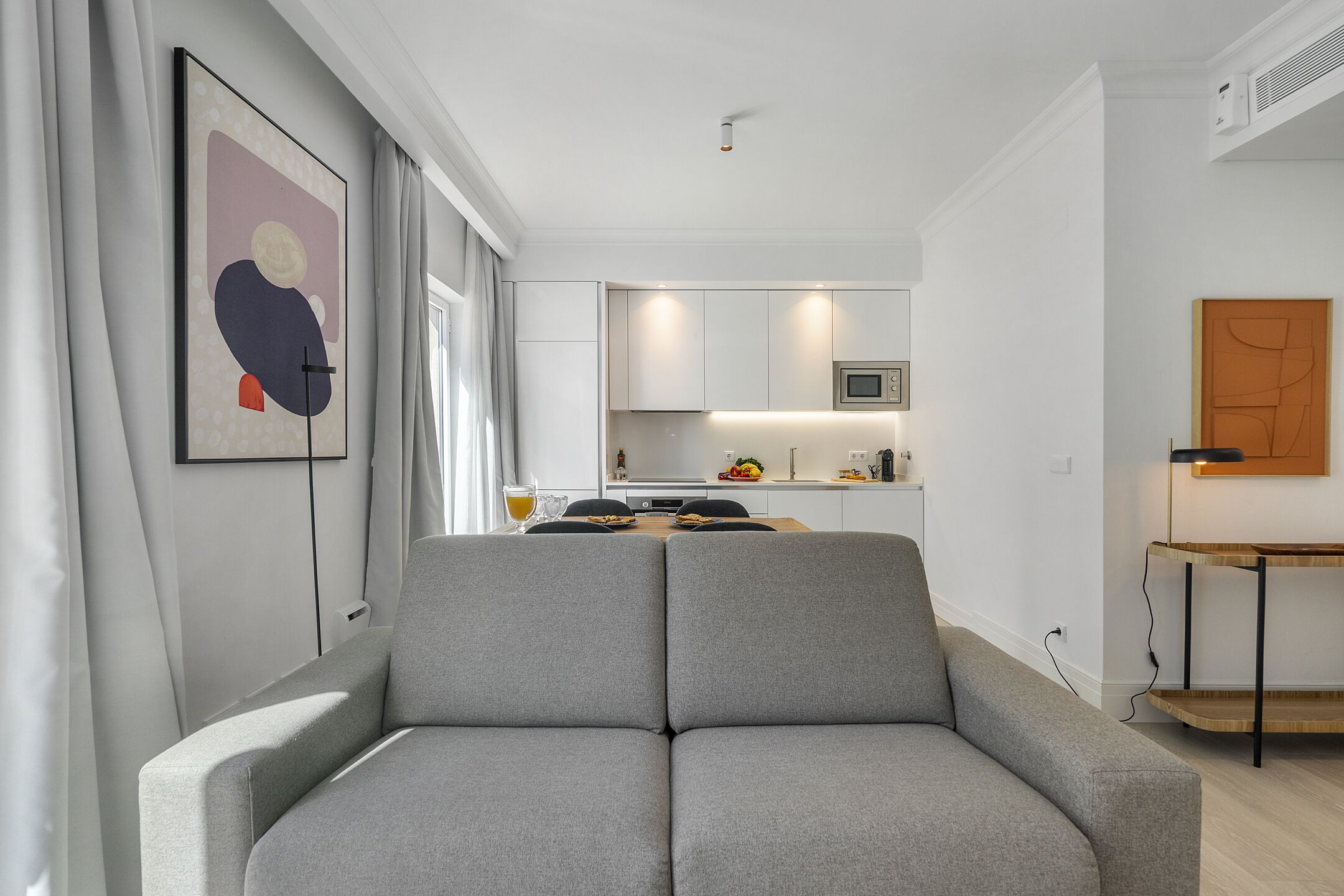 Uma sala de estar moderna com um sofá cinzento, uma área de jantar com mesa e cadeiras, uma cozinha com armários brancos e uma pintura abstrata emoldurada na parede.
