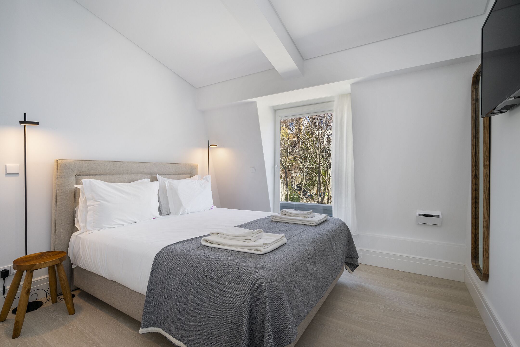 Une chambre à coucher moderne avec un lit, des lampes de chevet, une fenêtre donnant sur un beau paysage et un petit tabouret.