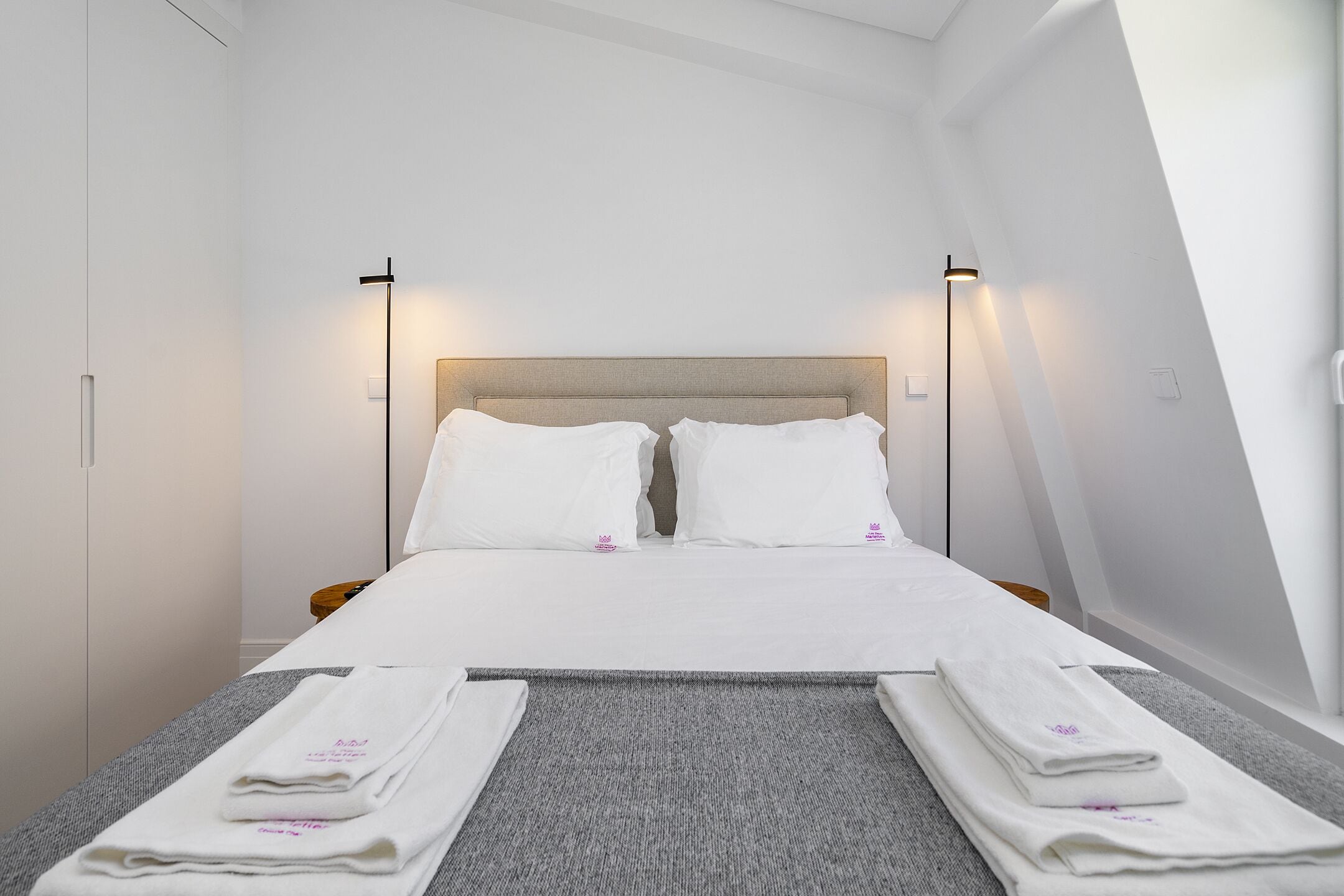 A neatly made bed with white linens and two pillows, a grey carpeted floor, and two bedside lamps.
