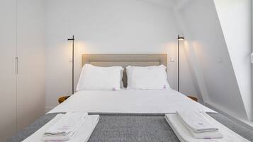 A neatly made bed with white linens and two pillows, a grey carpeted floor, and two bedside lamps.