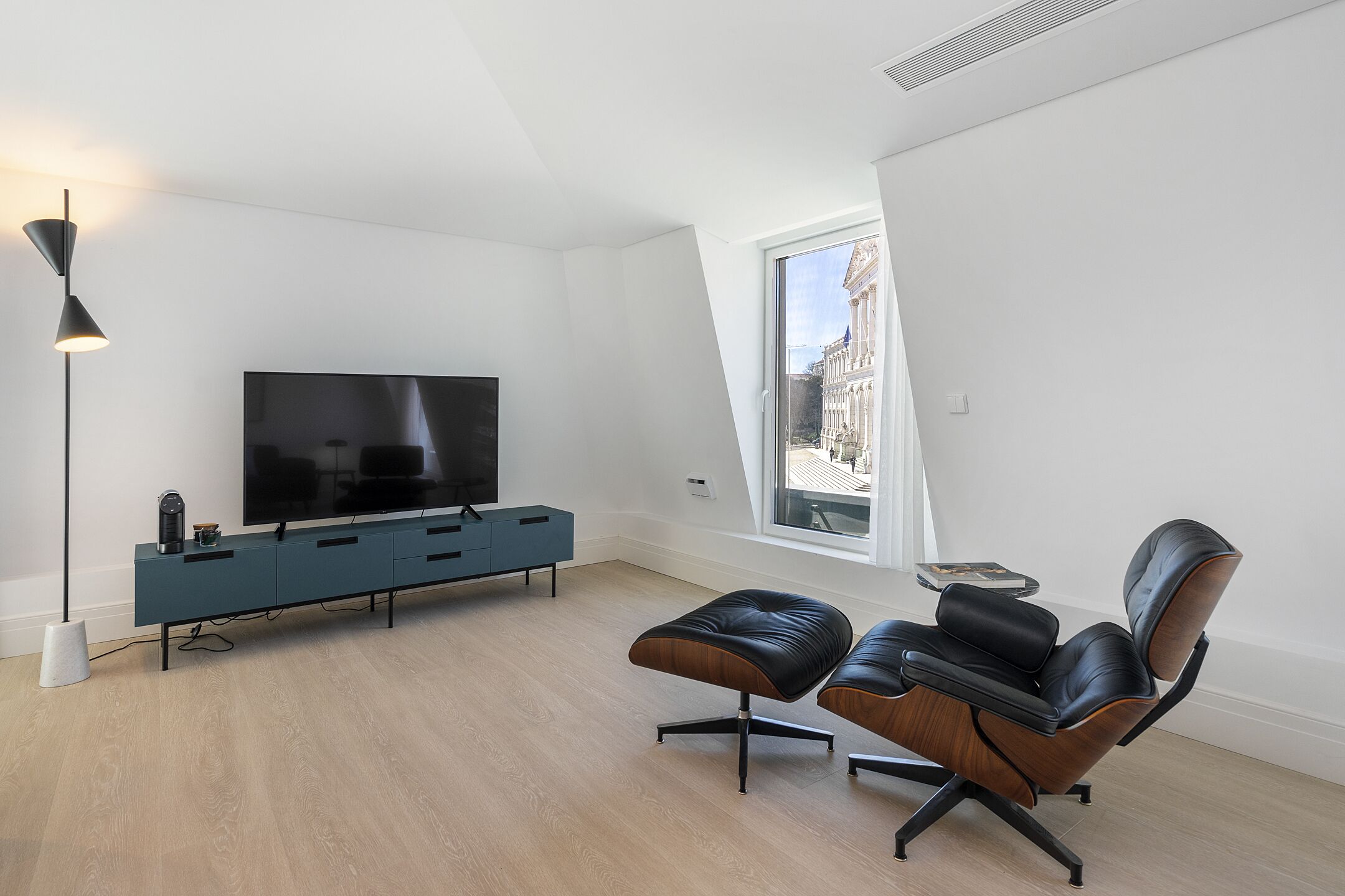 A modern living room with a flat-screen TV, a blue console, leather chairs, and a large window with a view of a building.