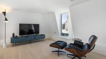 A modern living room with a flat-screen TV, a blue console, leather chairs, and a large window with a view of a building.