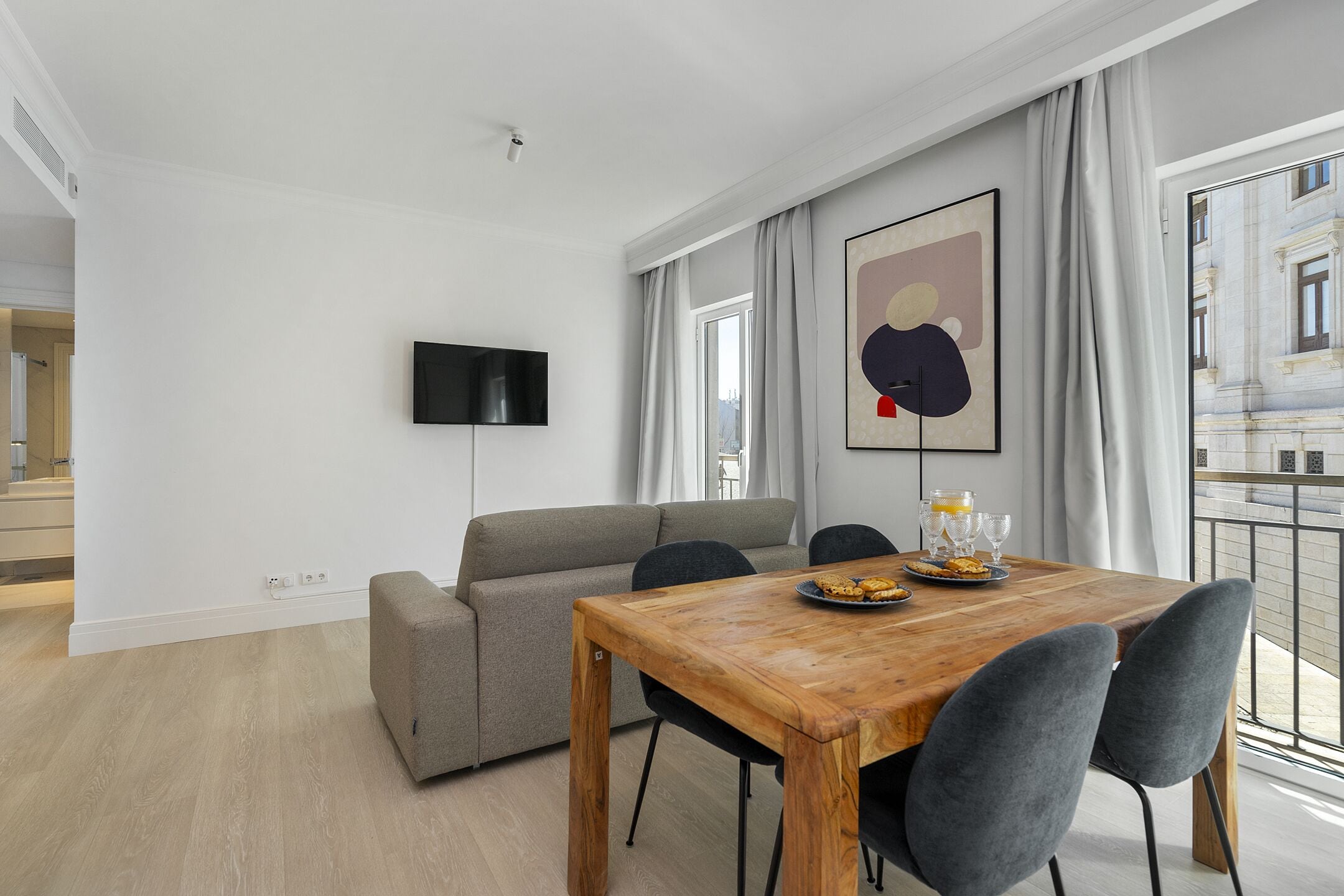 A modern living room with a wooden dining table, grey sofa, and a flat-screen TV mounted on the wall.