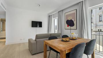 A modern living room with a wooden dining table, grey sofa, and a flat-screen TV mounted on the wall.