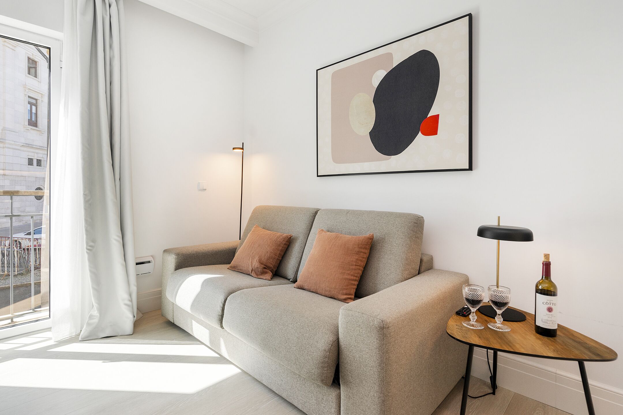 A modern living room with a grey sofa, two brown cushions, a wooden side table with a bottle of wine and two glasses, and a floor lamp.