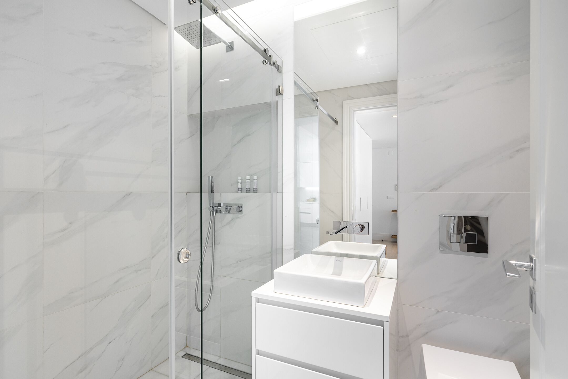 Un baño moderno con ducha de vidrio, paredes de mármol blanco y un tocador doble.