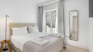 A hotel room with a large bed, a bedside table, a lamp, a window with curtains, and a mirror.