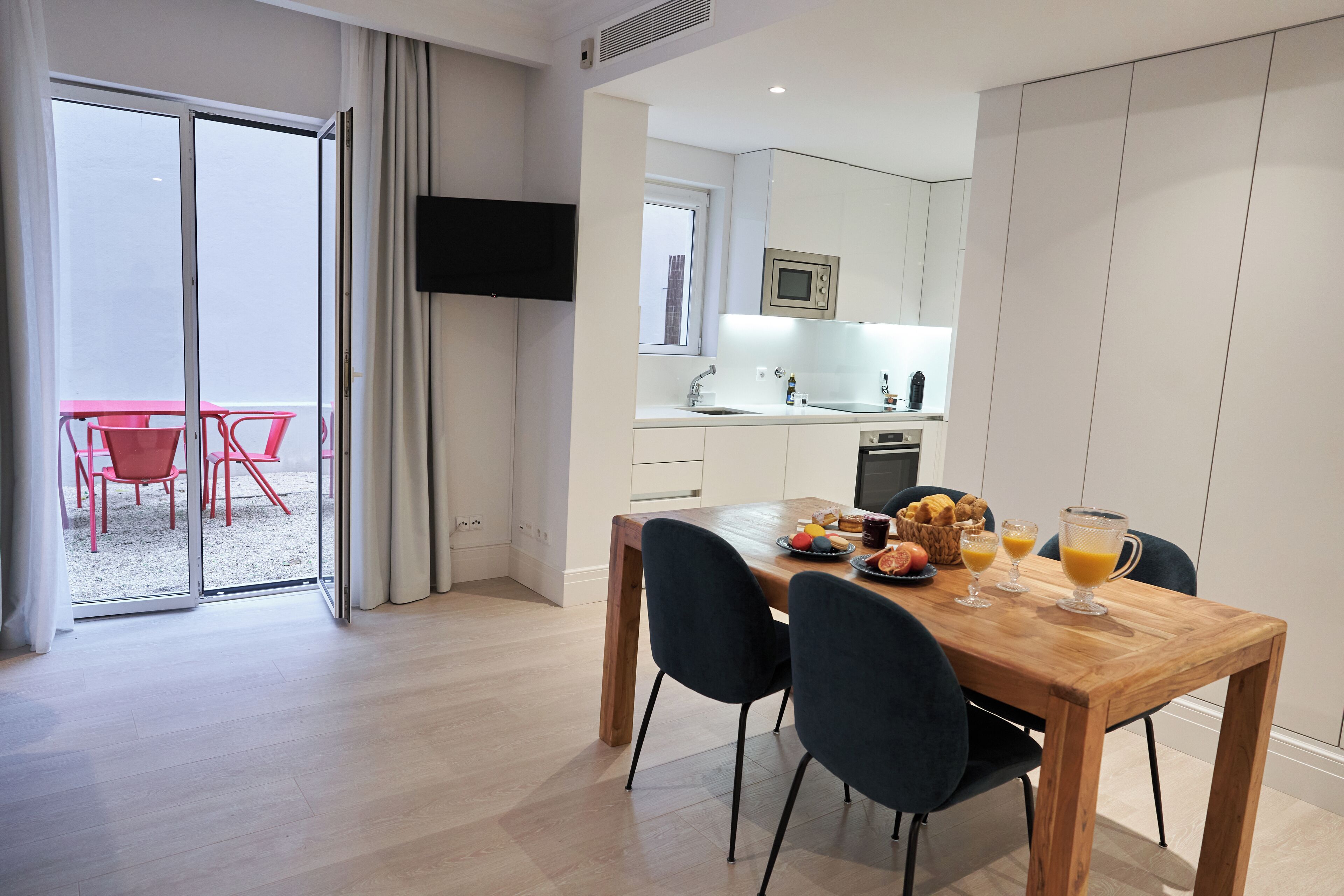 Eine moderne Küche mit Essbereich, einem über der Arbeitsfläche montierten Fernseher und Blick auf einen Balkon mit roten Stühlen.