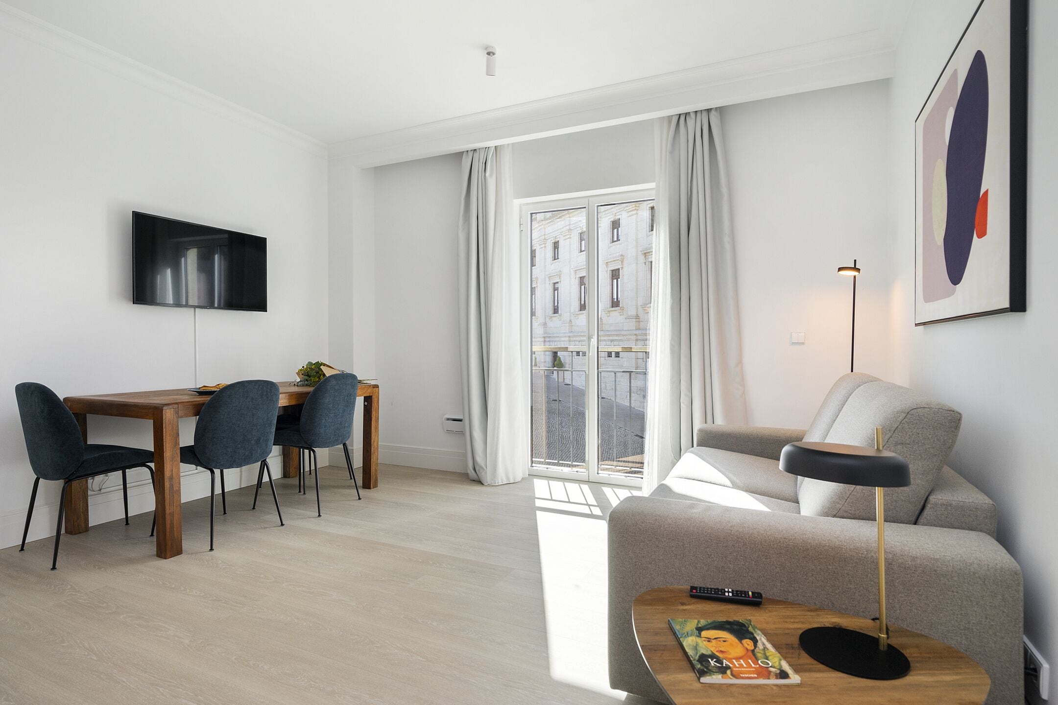 Una sala de estar moderna con sofá, una mesa de comedor con sillas, un televisor montado en la pared y un amplio ventanal con cortinas.