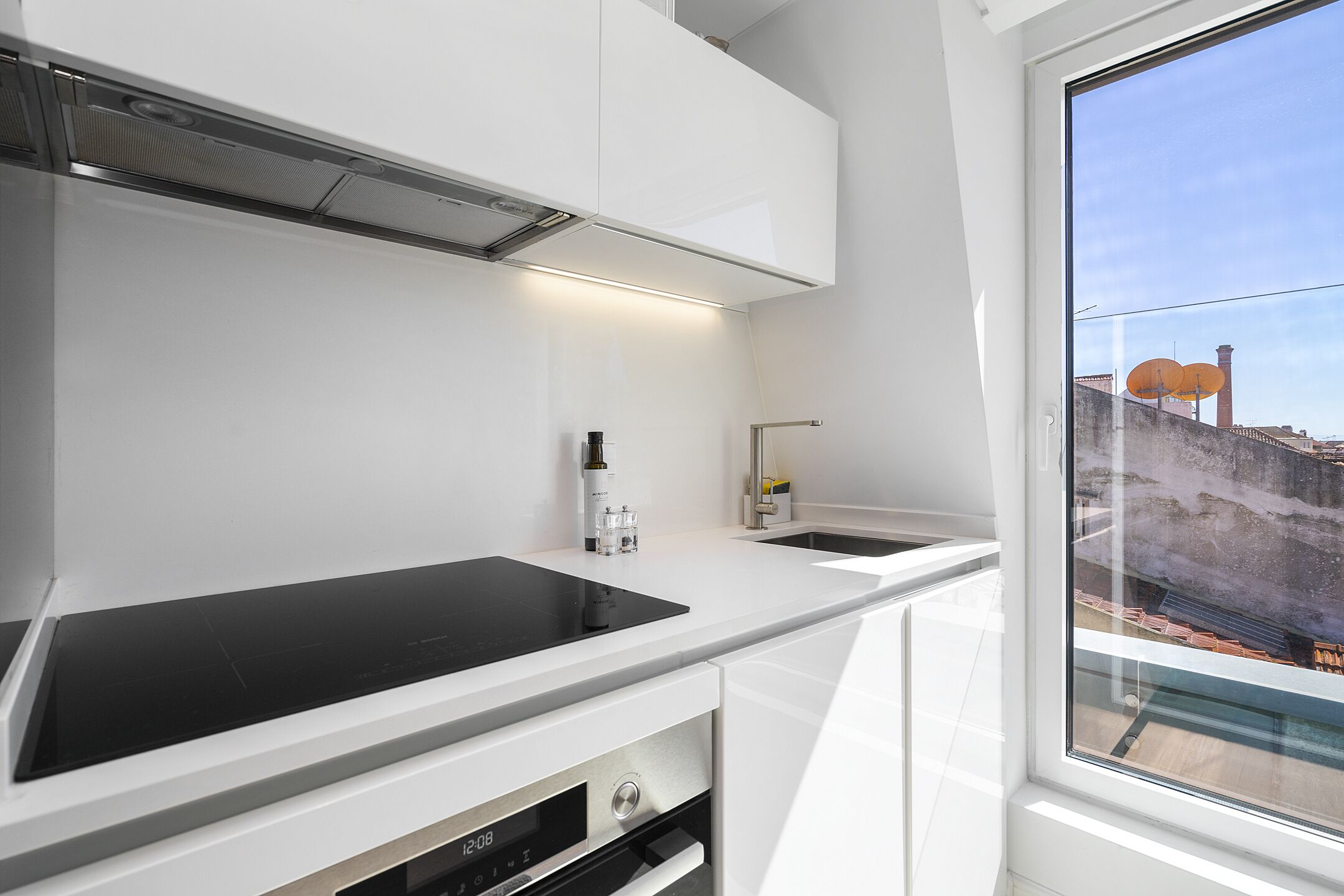 A modern kitchen with white cabinetry, a black induction cooktop, and a window with a view of buildings and a chimney.