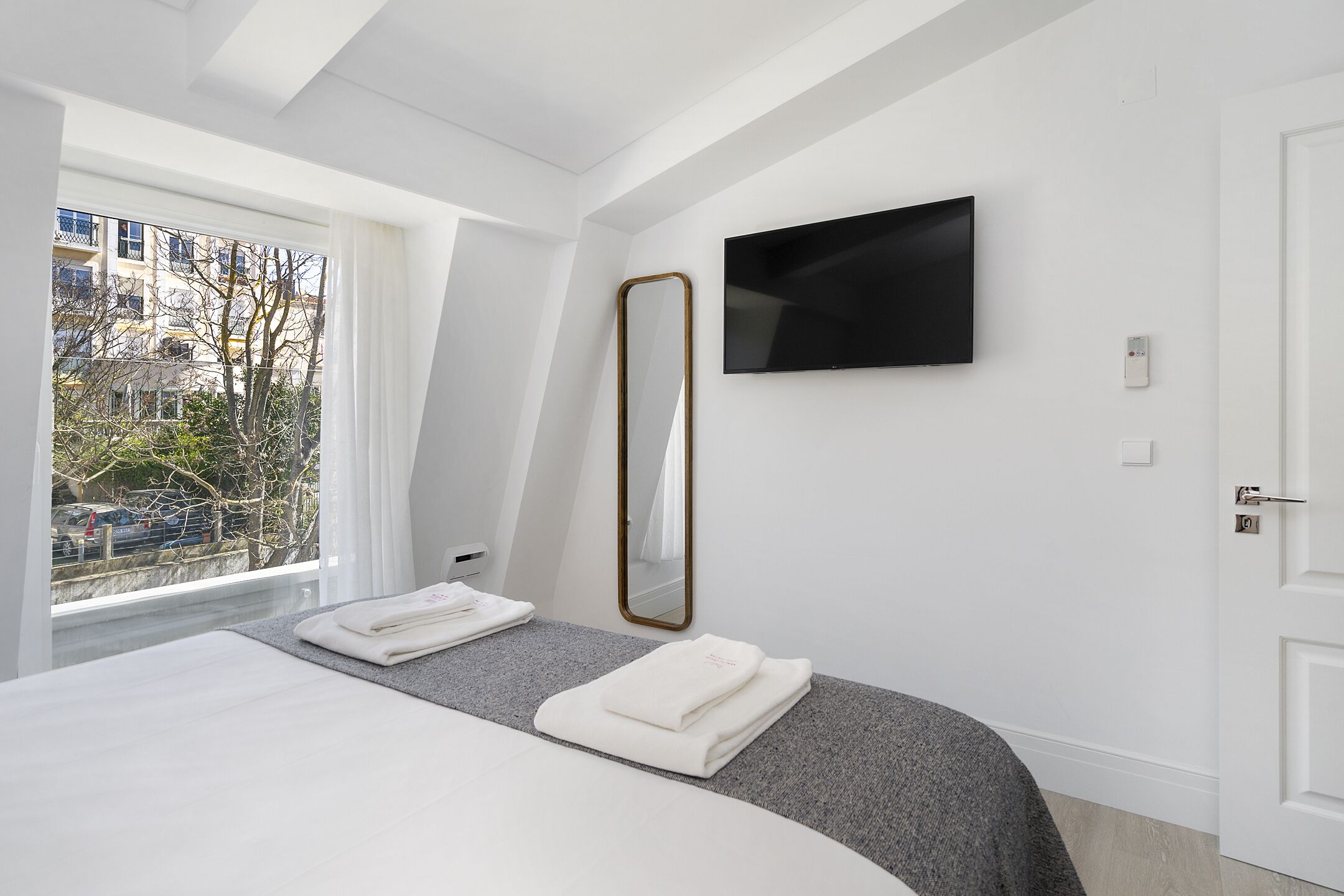 A modern hotel room with a bed, a large mirror, a flat-screen TV, and a window with a view of buildings and trees.