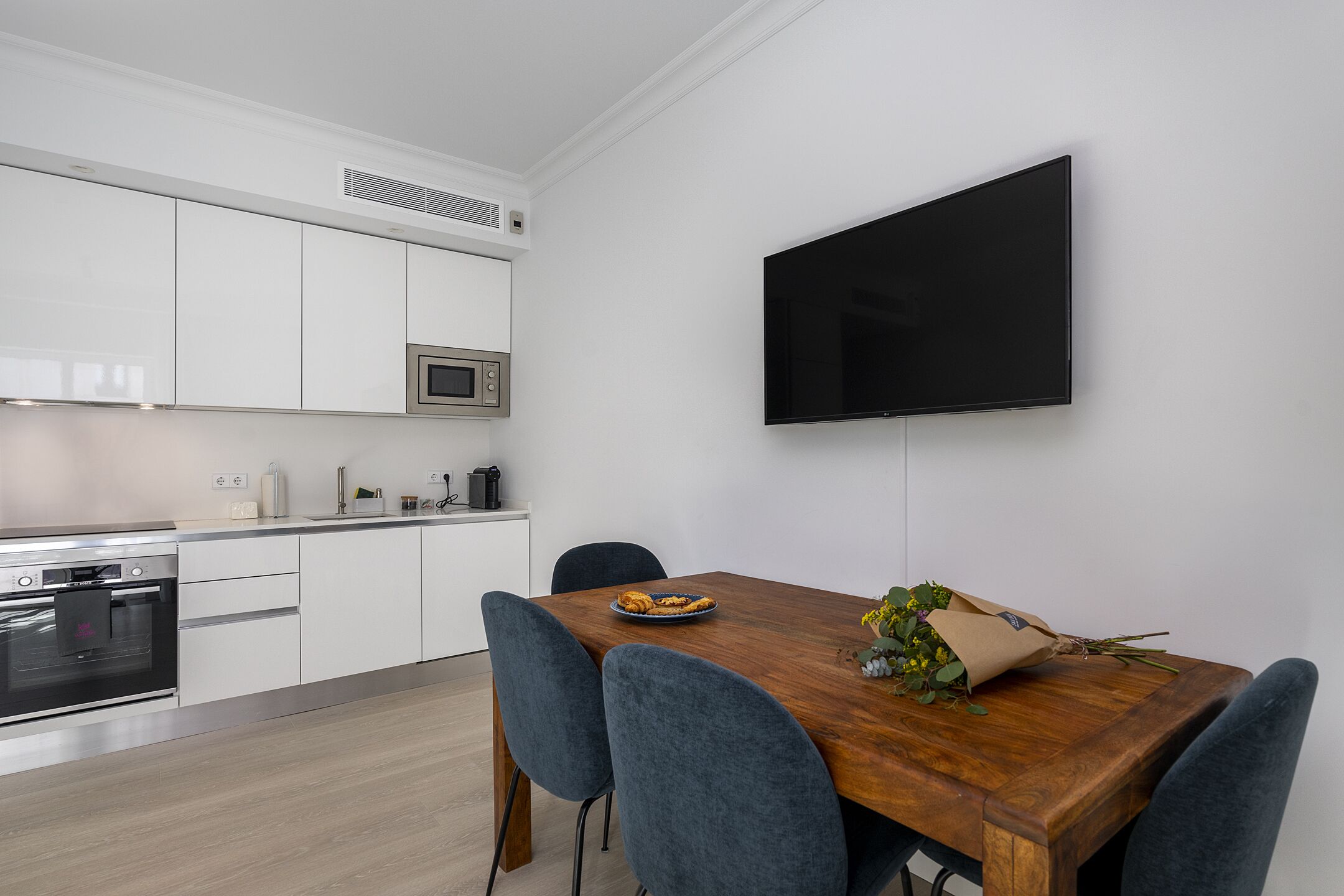 A modern kitchen with white cabinets, a built-in oven, and a microwave. There is a wooden dining table with chairs and a bouquet of flowers on it.