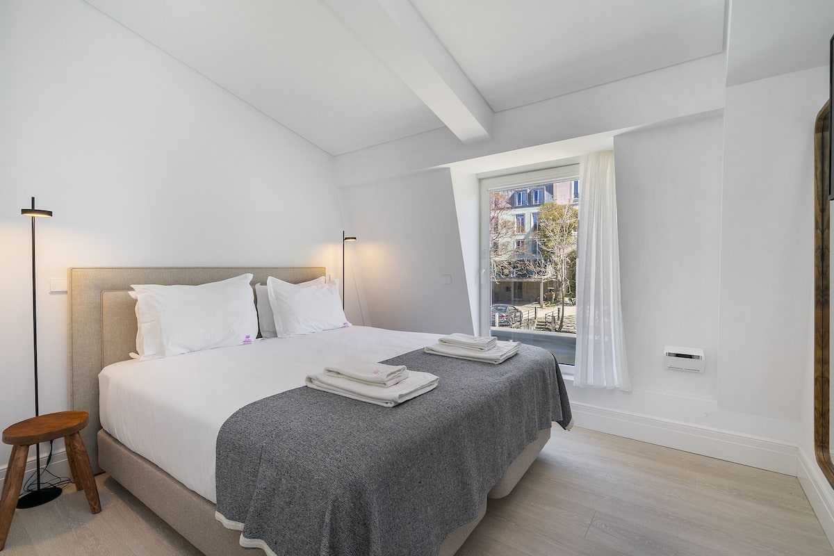 Una camera da letto moderna con un letto grande, uno sgabello, una finestra con tende e vista sugli edifici.