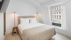 A bedroom with a bed, a TV mounted on the wall, a window with a view of buildings, and a floor lamp.