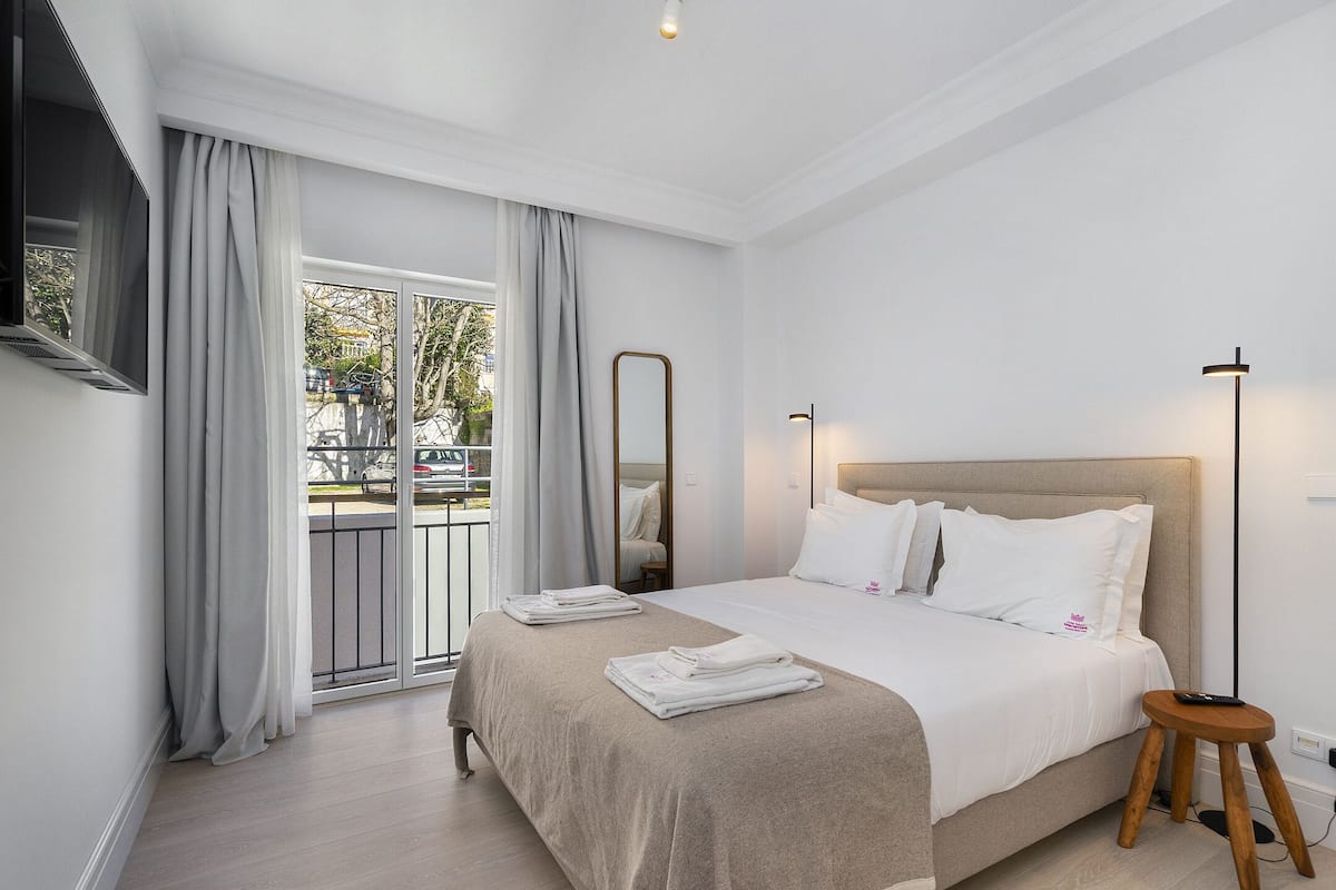 Una camera d'albergo con un letto, uno sgabello di legno, una televisione a schermo piatto e un'ampia finestra con tende.