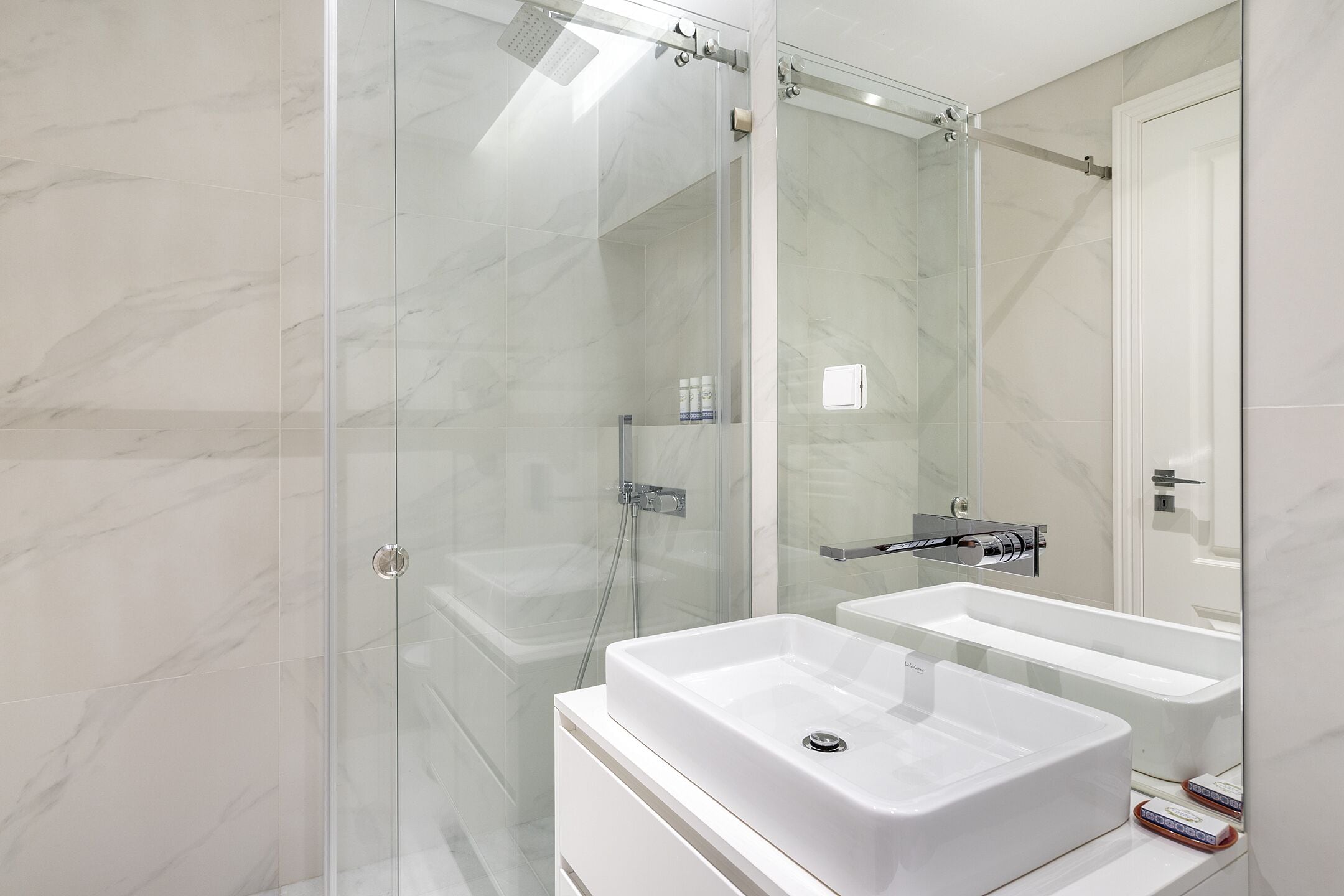 Un baño moderno con ducha envuelta en vidrio, lavamanos blanco y un espejo grande.