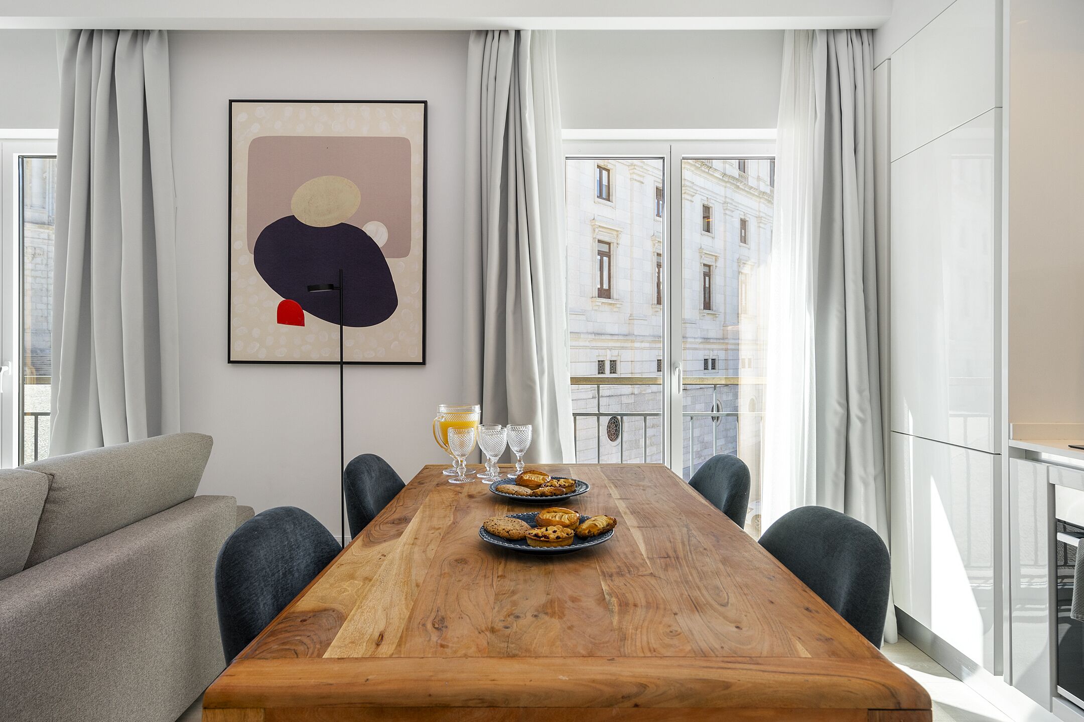 Un espace repas moderne avec une table en bois dressée pour deux personnes, un canapé et une vue sur la ville grâce à de grandes fenêtres.