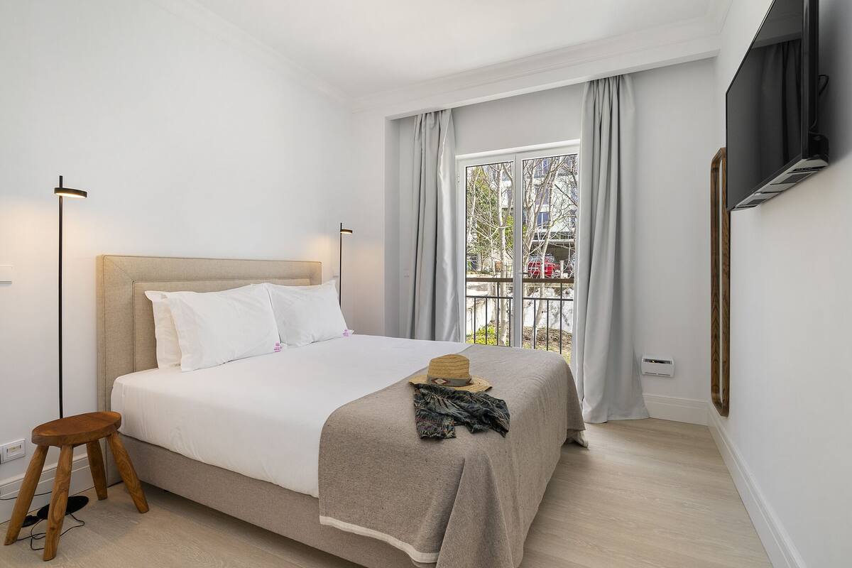 Un letto rifatto con cura, con una testiera in legno, un piccolo sgabello in legno e un cappello sul letto.
