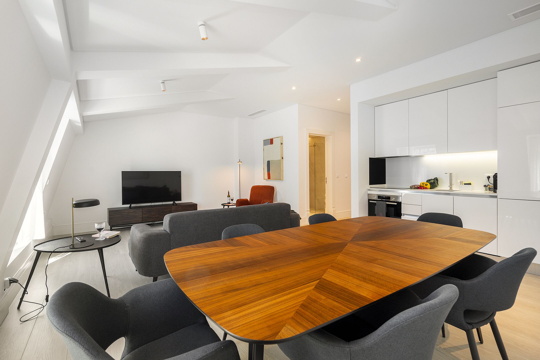 Ein modernes Wohnzimmer mit einem großen Esstisch aus Holz, grauen Sofas und einem Flachbildfernseher.