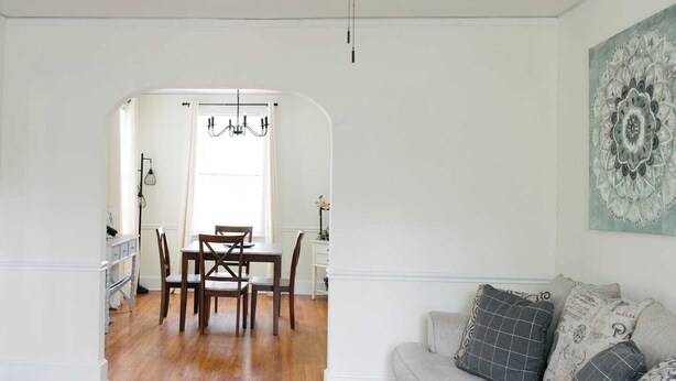 Un coin repas avec une table et des chaises en bois, un lustre et, à travers une arche, une vue sur la cuisine.