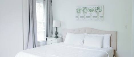 A bedroom with a bed, a nightstand, a lamp, a window with curtains, and a wall-mounted artwork.