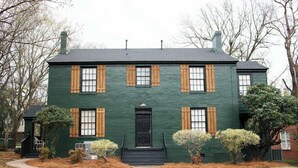 Une maison à deux étages, avec une façade verte et des volets en bois, une porte d’entrée noire et un petit porche à l’avant.