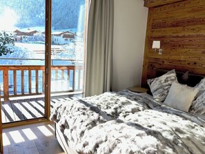 Une chambre à coucher avec une t ête de lit en bois, une grande fenêtre donnant sur des maisons enneigées et un couvre-lit blanc et moelleux.